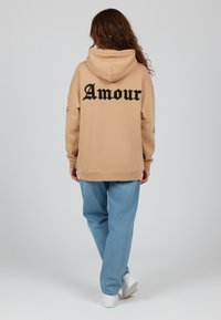 Felpa beige con "Amour" in grassetto nero sulla schiena, abbinata a pantaloni blu larghi e sneakers bianche, su uno sfondo neutro.