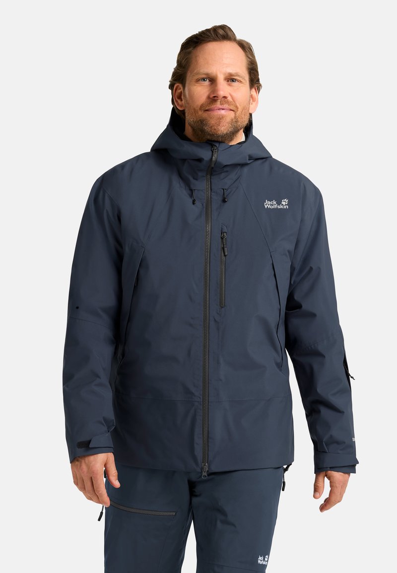 Giacca impermeabile navy con cappuccio, zip anteriore e numerose tasche con cerniera. Presenta polsini elastici e un logo sul lato sinistro del petto.