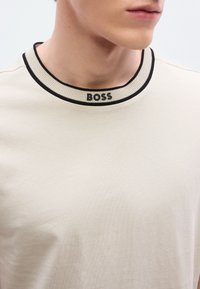 Μπεζ T-shirt με ριμπ κολάρο σε μαύρο και εκρού, με το λογότυπο "BOSS" να εμφανίζεται έντονα με μαύρα γράμματα στη λαιμόκοψη.