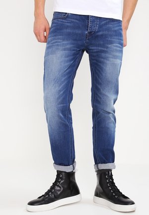 Muž ve vyhrnutých modrých džínách a černých kotníkových teniskách s bílou podrážkou stojí před jednobarevným pozadím.