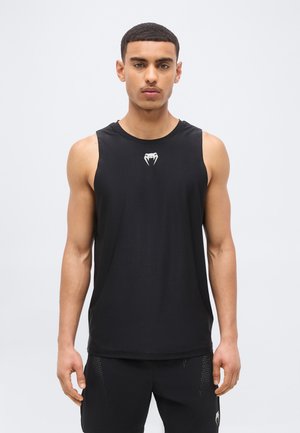 Junger Mann in schwarzem ärmellosem Sporttop und passenden Shorts, der vor einem schlichten hellen Hintergrund steht und nach vorne blickt.