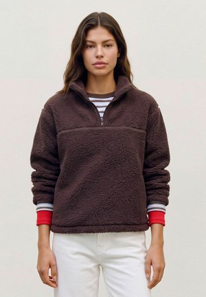 Modèle portant un pull en polaire marron foncé à demi-zip par-dessus une chemise à rayures et un pantalon blanc, sur un fond uni.