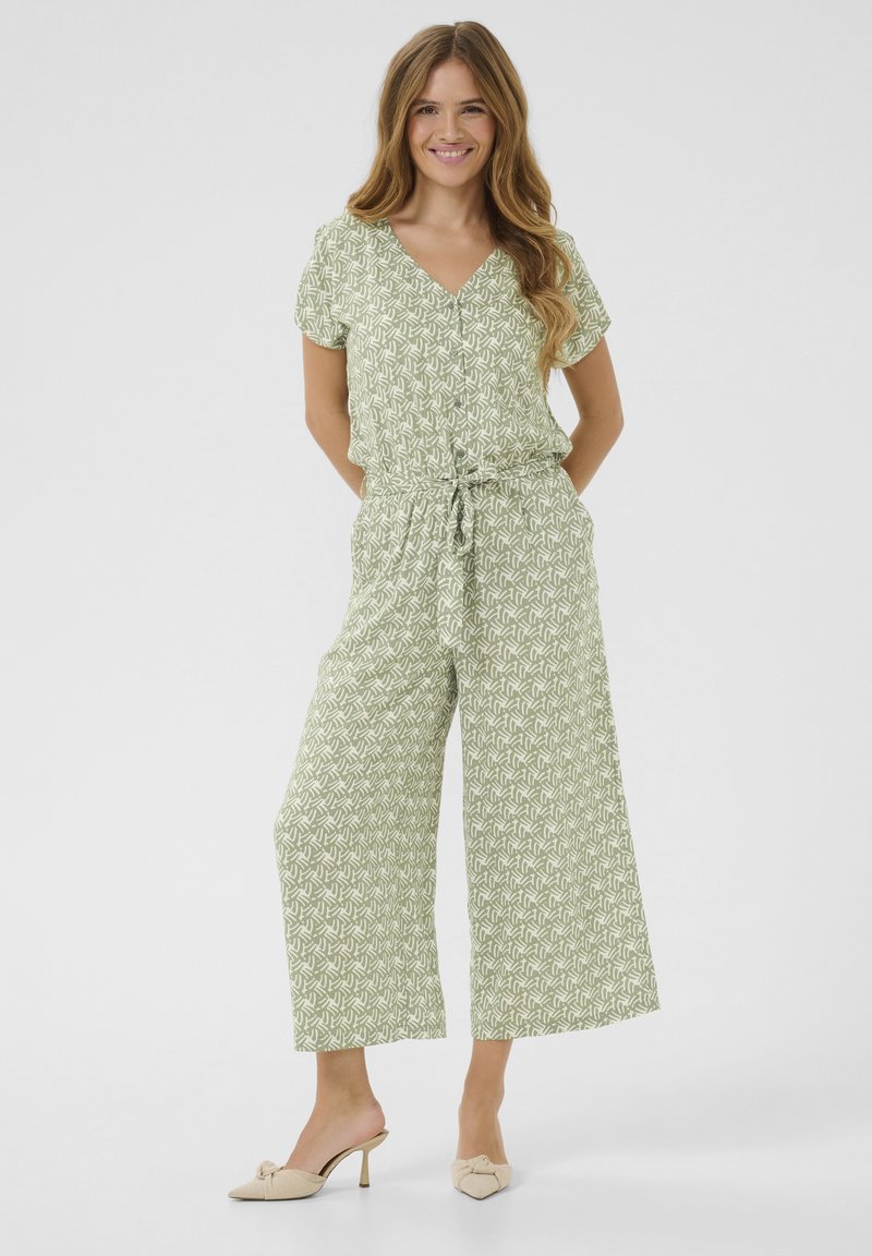 Combinaison verte à motifs avec des manches courtes, décolleté en V, taille à cordon, et jambes larges, associée à des mules à talons clairs.