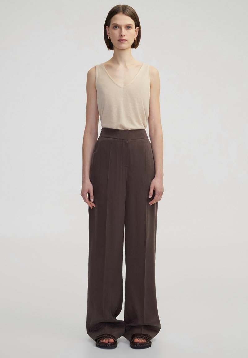 Krémový bezrukávový top s výstřihem do V, který je spárován s volnými, tmavohnědými kalhotami s vysokým pasem, s plisováním a měkkou látkou. Černé sandály.