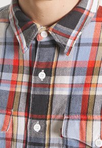 Geruit overhemd met knopen in rood, blauw, zwart en geel. Heeft een klassieke kraag en twee borstzakken met knopen. Zachte textuur.