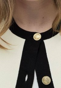 Detailní záběr na výstřih černobílé kardiganu se dvěma velkými zlatými dekorativními knoflíky a prameny blond vlasů na levé straně.