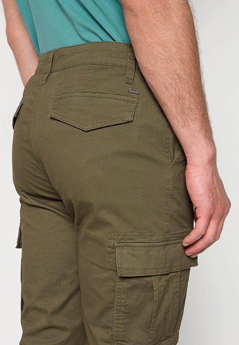 Mężczyzna ubrany w oliwkowe spodnie bojówki z kieszeniami z patkami i turkusową koszulę, stojący z ręką przy udzie na neutralnym tle.