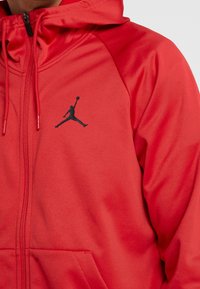 Sudadera con cremallera roja hecha de tela suave, que presenta un logo negro de Jumpman en el pecho y cordones en la capucha.