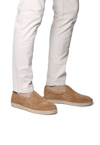 Des chaussures slip-on en suède tan avec des accents rayés et une semelle en jute de style espadrille. Portées avec un pantalon blanc ajusté.