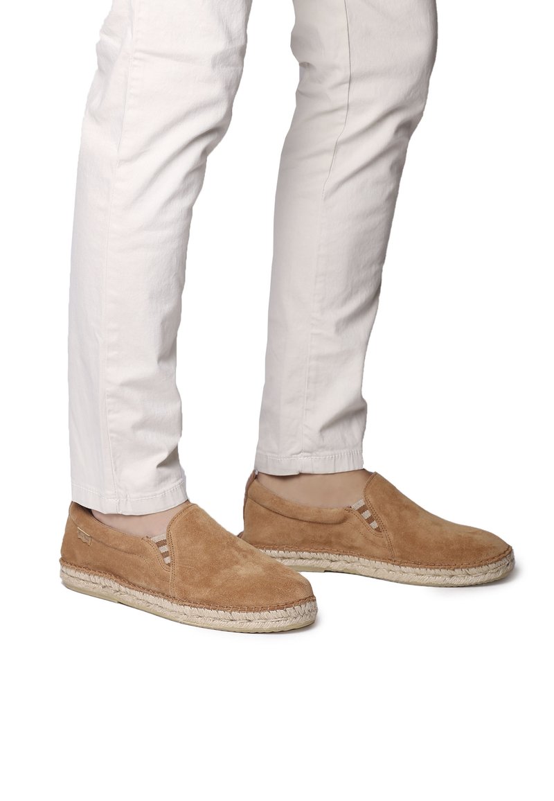 Des chaussures slip-on en suède tan avec des accents rayés et une semelle en jute de style espadrille. Portées avec un pantalon blanc ajusté.
