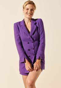 Robe blazer double boutonnage violette en tissu texturé, dotée de grands boutons et d'une coupe cintrée, associée à un short assorti.