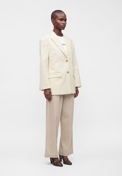 Femme aux cheveux courts portant un blazer crème oversize, un pantalon large beige et des chaussures à talons hauts marron foncé, debout sur un fond blanc.
