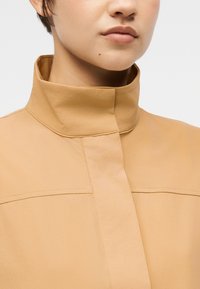 Κοντινό πλάνο ατόμου που φοράει μπεζ μπουφάν με ψηλό γιακά και κρυφή εμπρόσθια κλείστρα, μπροστά σε απλό φωτεινό φόντο.