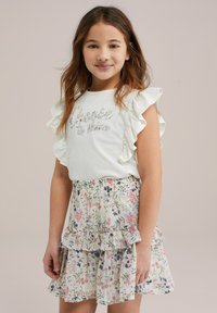 Balta trumpa rankovėmis marškinėliai su voliūriniais kraštais ir tekstu "Pasirink švytėti", derinami su sluoksniuota gėlių rašto sijonu pastelinių spalvų.