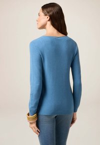 Pull en tricot bleu avec des manches longues, encolure dégagée et ourlet côtelé. Le modèle porte des bracelets empilés au poignet, assorti à un jean en denim.