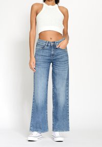 Žena v bílém pleteném crop topu, s vysokým pasem modrými širokými džíny a bílými platformovými teniskami, stojící s jednou rukou v kapse.