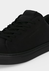 Κοντινό ενός μαύρου sneakers χαμηλού προφίλ με μαύρα κορδόνια και υφασμένη δερμάτινη γλωττίδα σε απλό φόντο.