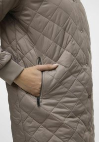 Gequilte, taupe buitenkleding met een ritszak aan de zijkant en ribgebreide boorden, gemaakt van een glad, lichtgewicht materiaal met een zachte glans.