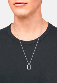 Zilveren ketting met een rechthoekige hanger, gedragen aan een fijne ketting, boven een zwart T-shirt met ronde hals. Strak ontwerp, gepolijste afwerking.