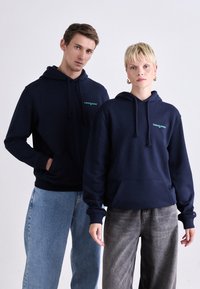 Sweatshirts bleu marine avec poches frontales et cordons, arborant un petit logo "tommy jeans" en turquoise. Portés avec un pantalon décontracté.