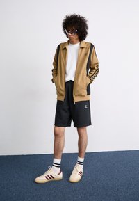 Tan zip-up jacket with black striped accents, paired with black shorts. White t-shirt underneath and cream sneakers with maroon stripes.