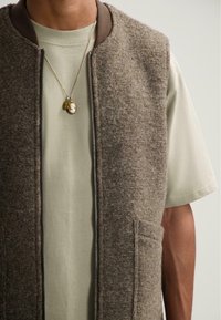 Muži v béžové košili s krátkým rukávem a hnědé texturované zapínací vestě se dvěma závěsnými náhrdelníky.