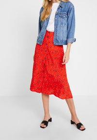 Jupe midi rouge à fleurs avec boutons à l'avant, en tissu léger ; associée à une veste en denim bleu clair et des sandales à talons carrés noires.