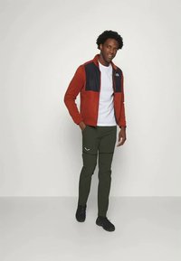 Červeno-námornická zipová bunda s texturovanou tkaninou, v kombinaci se zelenými kalhotami s bílým logem. Model stojí na neutrálním pozadí.