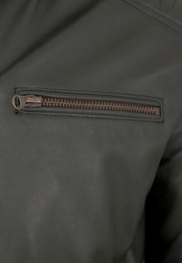 Schwarze Lederjacke mit diagonal platziertem, bronzefarbenem Reißverschlussfach mit strukturierten Details. Glatte Oberflächenstruktur mit dezentem Glanz.