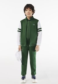 Zeleni sportski trenirka s bijelim detaljima i prugama. Sadrži jaknu na zip, polo ovratnik i logo, uparen s odgovarajućim hlačama i tenisicama.