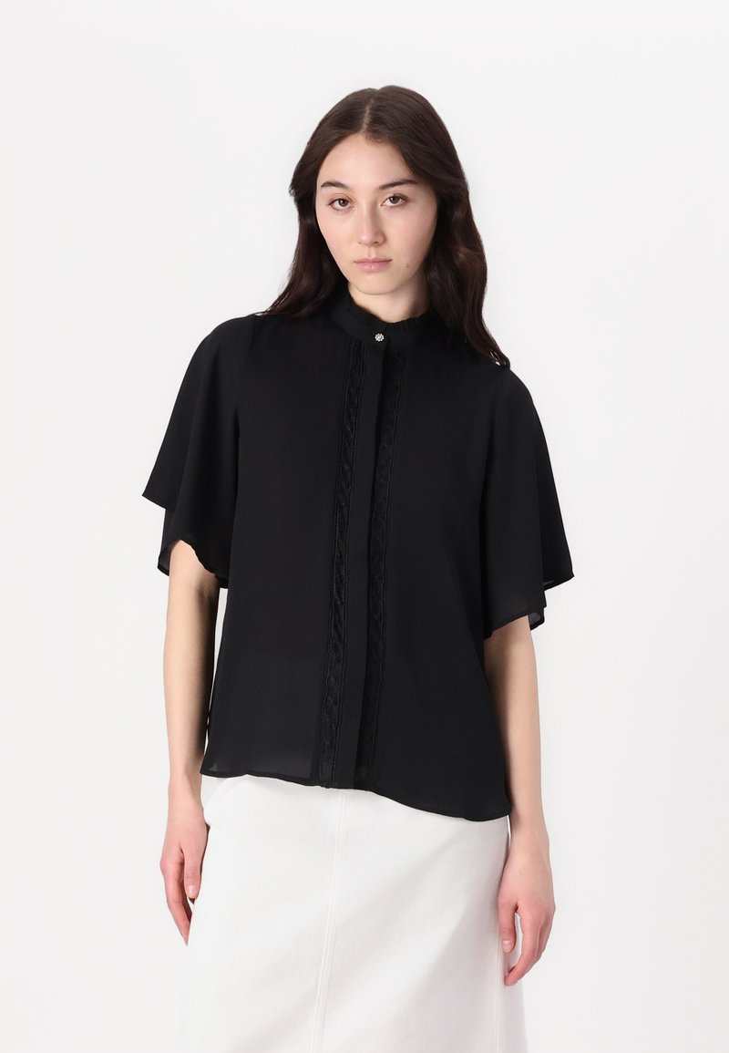 Blusa nera realizzata in tessuto leggero, con colletto, chiusura anteriore con bottoni e dettagli decorativi in pizzo verticale. Maniche a sbuffo corte.
