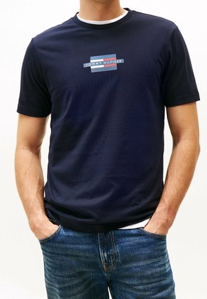 Mężczyzna ubrany w granatową koszulkę Tommy Hilfiger z logo na środku klatki piersiowej i niebieskie dżinsy, ręce w kieszeniach.