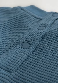 Modrý tkaný materiál s textúrou vaflí a zapínaním na gombíky. Gombíky sú okrúhle a matné, čo dodáva funkčný detail dizajnu.