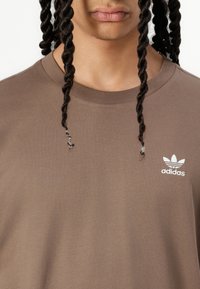 Brūns kokvilnas T-krekls ar apaļu kaklu un īsām piedurknēm. Uz kreisās krūts zonas ir balta izšūta Adidas logo.