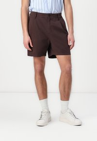 Braune Shorts mit einem elastischen Bund, zwei Seitentaschen und einem einzelnen Frontknopf. Getragen mit einem hellblauen gestreiften Hemd und weißen Sneakers.