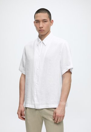 Ung mann med kort hår iført en hvit kortermet skjorte og beige bukser, står foran en ensfarget lys bakgrunn.