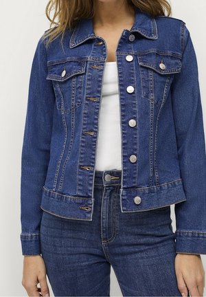 Femme portant une veste en jean bleu foncé avec des boutons argentés sur un haut blanc et un jean en denim bleu foncé assorti, debout les bras détendus.