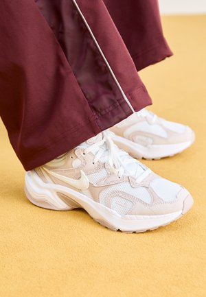 Baskets Nike blanches et beiges à lacets, portées avec un pantalon bordeaux sur une surface jaune, chaussures tournées vers la droite.