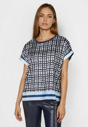 Woman with straight brown hair wearing a navy blue and white patterned short-sleeve top and dark blue leather pants against a plain background.