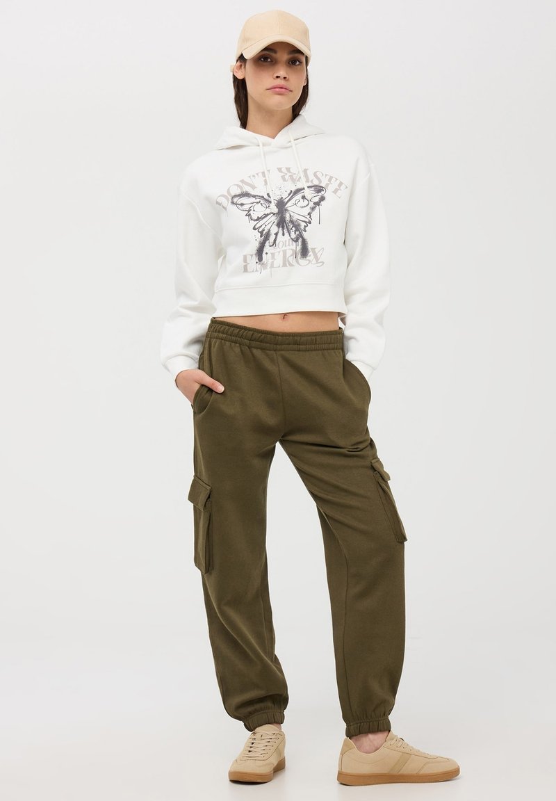 Obcisła biała bluza z kapturem z grafiką motyla, oliwkowe dresy z bocznymi kieszeniami i beżowe trampki. Gładkie tło.