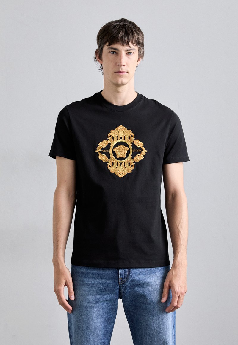 T-shirt en coton noir avec un design graphique brodé en or au centre, représentant des motifs ornés avec un motif de lion.