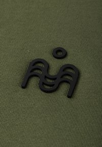 Musta abstrakti geometrinen muoto, jossa on kaarevia ja suoria viivoja sekä erillinen pieni ympyrä, sijoitettuna oliivinvihreälle, poikittain uurretulle kankaalle.