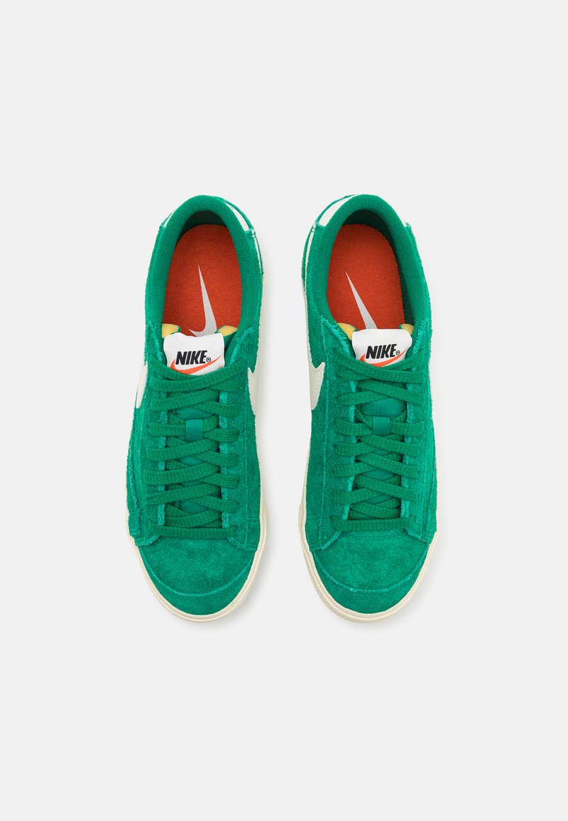 Zapatillas de ante verde con branding blanco, puntera redonda y cordones planos. Acentos rojos y amarillos visibles en el interior. Diseño clásico de corte bajo.