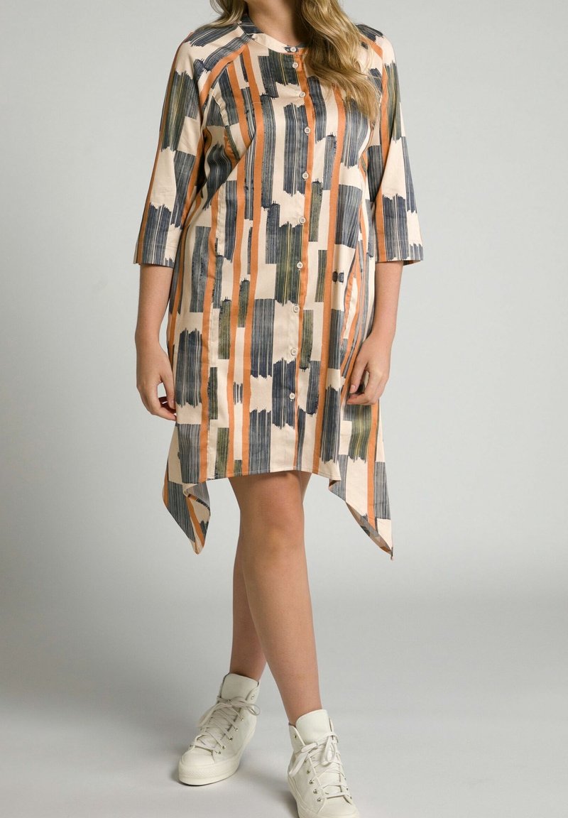 Femme portant une robe boutonnée mi-longue aux genoux avec des rayures verticales en coups de pinceau orange, noir et vert, et des baskets blanches montantes.
