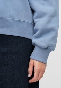 Felpa di cotone azzurro chiaro con polsini a coste, dettagli delle cuciture visibili, abbinata a un tessuto scuro sottostante. Mano parzialmente visibile.