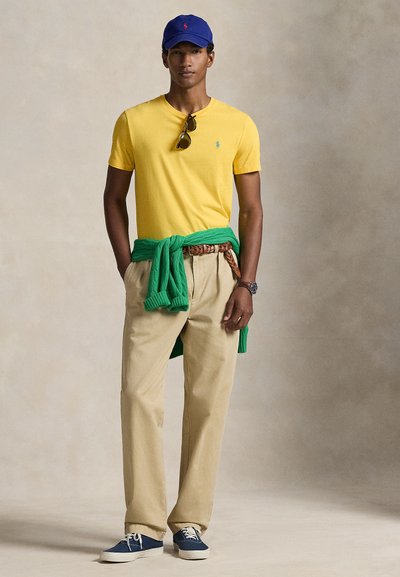 Κίτρινη βαμβακερή μπλούζα, πράσινο πουλόβερ δεμένο γύρω από τη μέση, μπεζ παντελόνι και μπλε πάνινα παπούτσια. Αξεσουάρ με γυαλιά ηλίου και μπλε καπέλο.