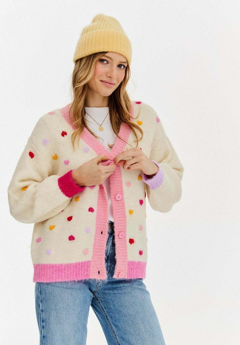 Kremowy kardigan z kolorowymi sercami, różowym i lawendowym wykończeniem oraz zapięciem na guziki. Noszony na białej bluzce z niebieskimi dżinsami. Żółta dziana czapka.