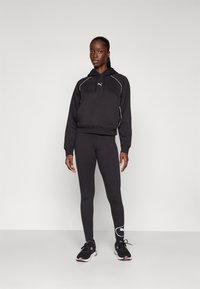 Svart kortermet hettegenser med hvit piping og logo, kombinert med tettsittende svarte leggings og svarte joggesko, med lagedetalj på skoen.