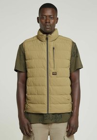 Chaleco tipo puff acolchado color beige con cuello alto, cierre de cremallera y bolsillo en el pecho. Lleva una camisa de patrón verde oscuro debajo. Acabado mate.