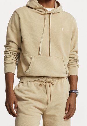 Persona che indossa una felpa beige con cappuccio e pantaloni della tuta abbinati, con un orologio al polso con cinturino blu navy etichettato "POLO".